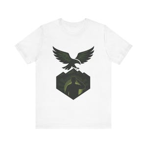 May include: A white short-sleeve t-shirt featuring a graphic print of a dark green eagle in flight above a hexagonal emblem. The emblem contains a silhouette of a person saluting against a backdrop of mountains and a circular sunburst design.