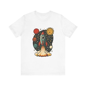 以下が含まれることがあります： 宇宙へ飛び立つロケットが中央に描かれた白い半袖Tシャツ。デザインには、笑顔の太陽、3つの惑星、星が暗い背景に配置されており、ロケットの下からはオレンジと黄色の煙が立ち上っています。