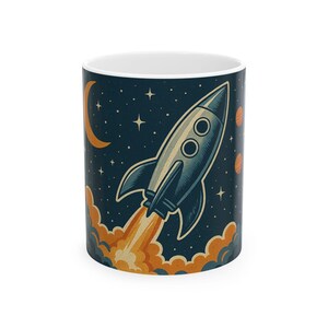 May include: A cylindrical ceramic mug featuring a dark navy blue background with a graphic illustration of a silver rocket ship launching into space. The design includes orange clouds of smoke, a crescent moon, stars, and two small orange planets.