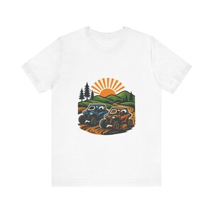 Op de afbeelding: Een wit T-shirt met korte mouwen voorzien van een grafische illustratie van twee terreinwagens die op een zandpad rijden door een landschap met groene heuvels, dennenbomen en een heldere oranje zon die opkomt op de achtergrond.
