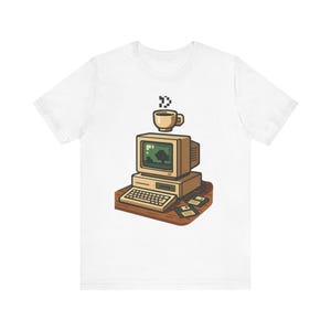 Pode incluir: Uma t-shirt branca com um gráfico de um computador vintage bege sobre uma secretária castanha. A ilustração inclui um monitor, um teclado, duas disquetes e uma chávena de café a fumegar acima do ecrã, tudo em estilo pixel art.