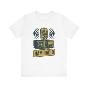 May include: A white short-sleeve t-shirt featuring a graphic print of a vintage-style microphone positioned in front of a radio transceiver. The design includes dark blue and yellow colors with the text HAM RADIO printed on a rectangular sign.