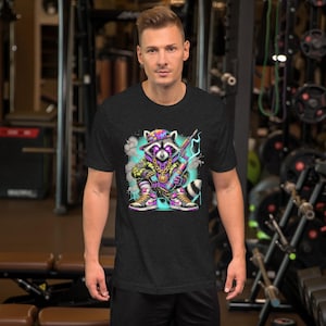 May include: Dark gray t-shirt featuring a colorful graphic of a raccoon wearing a hat, gold chain, and holding a weapon. The raccoon is depicted in a graffiti-style design with purple, yellow, and blue accents. The shirt is short-sleeved.