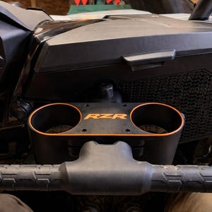Puede incluir: Portavasos RZR negro y naranja con dos aberturas circulares. El logotipo de RZR es naranja. El portavasos está montado en un salpicadero negro con un manillar negro debajo.
