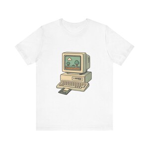 May include: A white t-shirt featuring a graphic illustration of a vintage beige desktop computer with a keyboard and a floppy disk inserted. The computer screen displays a simple pixelated landscape with green trees, a blue sky, and a white cloud.