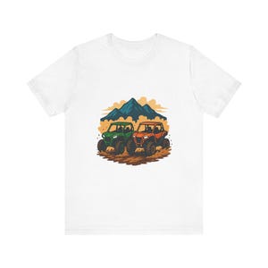 Może przedstawiać: Biały t-shirt z grafiką przedstawiającą zielony i pomarańczowy pojazd terenowy jadący po polnej drodze. W tle znajdują się niebieskie góry i żółte chmury, tworzące scenę przygody na świeżym powietrzu na przodzie koszulki.