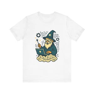 Può includere: Una t-shirt bianca con un'illustrazione grafica di un mago con barba lunga e cappello a punta, che tiene una bacchetta magica mentre usa un computer portatile. Il design include motivi di circuiti, ingranaggi e una nuvola di cifre binarie.