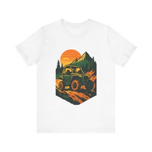 Może przedstawiać: Biały t-shirt z krótkim rękawem z nadrukiem graficznym przedstawiającym zielony pojazd terenowy jadący polną drogą. W tle znajduje się duże pomarańczowe słońce, ciemnozielone sosny i szczyt góry, wszystko zamknięte w sześciokątnym kształcie.