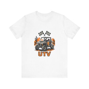 Könnte beinhalten: Ein weißes Kurzarm-T-Shirt mit einem Grafikdruck eines schwarzen UTV-Fahrzeugs in einer Wüstenlandschaft mit Kakteen, orangefarbenen Bergen und zwei karierten Rennflaggen. Der Text UTV ist in fetten orangefarbenen Buchstaben aufgedruckt.