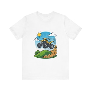 Peut inclure: Un t-shirt blanc avec une illustration graphique représentant un quad jaune sautant au-dessus d'une colline verte. Le motif comprend un soleil jaune vif, des nuages blancs et un nuage de poussière brune derrière les pneus sur un fond bleu.