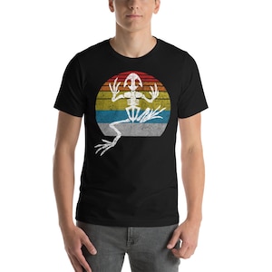 Puede incluir: Camiseta negra con un gráfico desgastado de un esqueleto de rana sobre un diseño de puesta de sol retro. El esqueleto de la rana es blanco, y la puesta de sol tiene bandas rojas, amarillas, azules y blancas. Una camiseta informal de inspiración vintage.