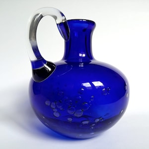 May include: A cobalt blue glass jug with a clear glass handle. The jug has a round body with a textured surface that resembles grapes.