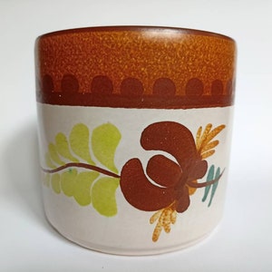 Könnte beinhalten: Eine braune und weiße Keramiktasse mit einem Blumendesign. Die Tasse hat einen braunen Rand und ein braunes Band mit einem sich wiederholenden Muster aus Kreisen.