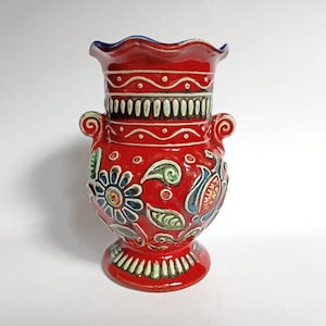 Puede incluir: Un jarrón de cerámica rojo con un diseño floral azul y blanco. El jarrón tiene un borde festoneado y dos asas.
