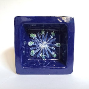 Puede incluir: Un pequeño plato cuadrado de cerámica azul con un diseño floral blanco y verde en relieve en el centro.