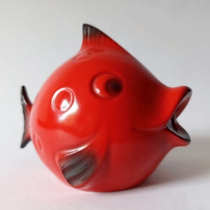 Può includere: Una statuetta di pesce in ceramica rosso vivo con dettagli neri. Il pesce ha un corpo grande e rotondo e una piccola bocca aperta.