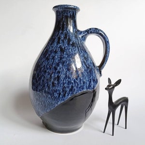 Peut inclure: Un vase en céramique bleu et noir avec un glaçage moucheté et un corps arrondi. Le vase a une anse et une ouverture étroite.
