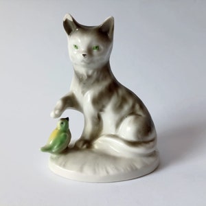 Puede incluir: Una figura de cerámica de un gato con marcas grises y blancas, ojos verdes y una pata levantada. Un pequeño pájaro amarillo y verde está sentado a los pies del gato. La figura está sobre una base blanca.