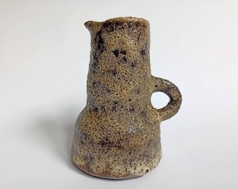May include: A small, brown ceramic pitcher with a textured surface and a single handle. The pitcher has a rounded base and a slightly flared top.