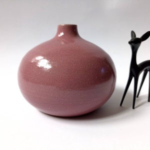 Può includere: Un piccolo vaso rotondo in ceramica rosa con una finitura liscia e lucida. Il vaso è su una superficie bianca.
