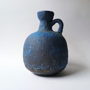 Può includere: Un vaso in ceramica blu e marrone con corpo arrotondato, manico e collo stretto. Il vaso ha una superficie strutturata e un colore blu scuro con accenti marroni. Il manico ha la forma di un anello.