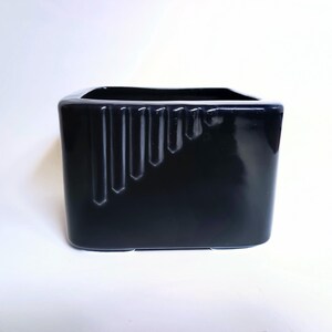 May include: A black ceramic vase with a rectangular shape and a glossy finish. The vase has a series of vertical, recessed lines on one side. The opening is square. The vase is displayed on a white surface.