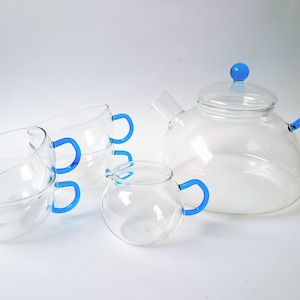 May include: A clear glass tea set with blue detailing. The set comprises a teapot with a blue handle and lid, a creamer, and three teacups with blue handles. The tea set is designed for serving tea.