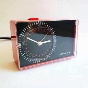 May include: A vintage, rectangular alarm clock with a red case and a black face. The clock face features white and red markings, with the text "electro boy" at the bottom. The clock has white hands and a red button on top.