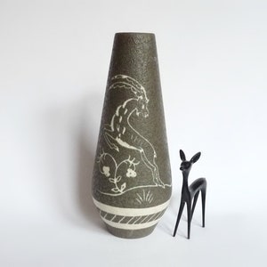 Op de afbeelding: Een hoge, groene keramische vaas met een wit ontwerp van een dier, mogelijk een steenbok, met een witte band rond de onderkant. De vaas staat op een wit oppervlak.