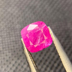 May include: A faceted, cushion-cut, hot pink gemstone is held by silver tweezers against a gray background. The gem's multiple facets reflect light, creating a brilliant display of color and sparkle. The gem is a vibrant, eye-catching piece.