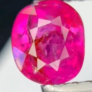Könnte beinhalten: Ein facettierter, oval geformter, pinkfarbener Edelstein, der von einer silbernen Pinzette gehalten wird. Der Edelstein hat ein sternförmiges Muster in der Mitte und ist hochreflektierend. Ein lebendiges, auffälliges Schmuckstück.