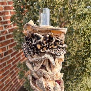 May include: A decorative bottle stopper with a layered design. The stopper features fabric with a leopard print and a cream-colored fabric with printed text. The stopper is designed to fit a bottle.
