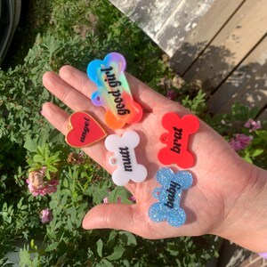 May include: A collection of five acrylic pet tags in various shapes and colours. One bone-shaped tag is multicoloured with the text good girl, a red heart tag says angel, a white bone tag says mutt, a red bone tag says brat, and a blue glitter bone tag says baby.