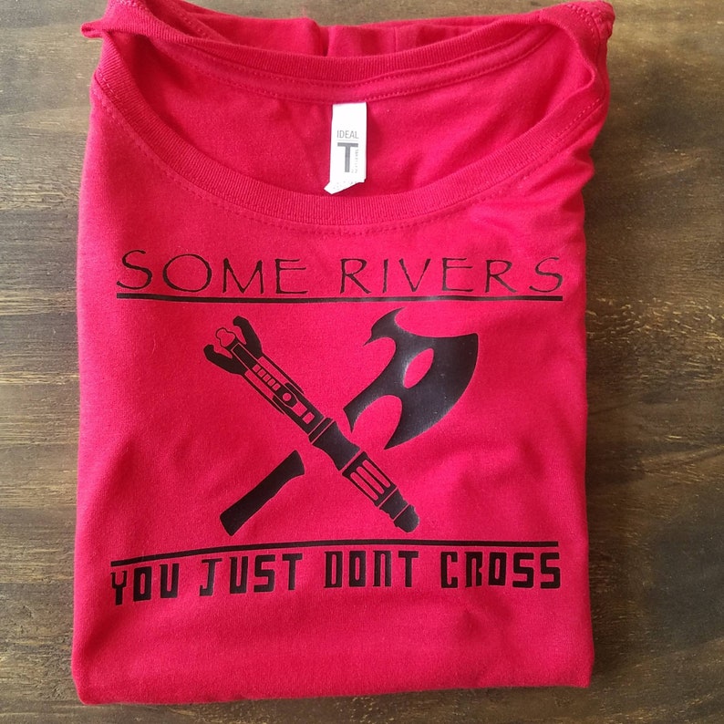 May include: A red t-shirt with black text and graphics. The text reads "SOME RIVERS YOU JUST DONT CROSS" above a crossed axe and a futuristic weapon. The shirt is folded on a wooden surface.