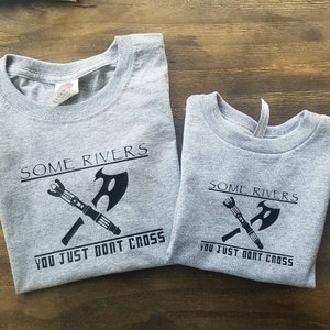 May include: Two gray t-shirts with the text "SOME RIVERS YOU JUST DONT CROSS" and a black graphic of a crossed axe and a futuristic weapon. The shirts are displayed on a wooden surface.