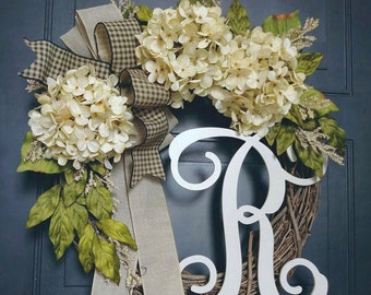 May include: A wreath made of grapevine with a white wooden letter R in the center. The wreath is decorated with white hydrangeas, green leaves, and a black and white checkered ribbon.