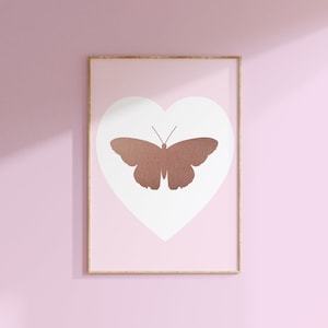 Puede incluir: Una impresión enmarcada con un fondo rosa y una forma de corazón blanca en el centro. Dentro del corazón hay una silueta de mariposa de color cobre.