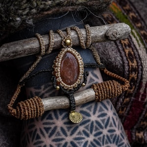 Op de afbeelding: Een handgemaakte macramé ketting met een druppelvormige, roodbruine stenen hanger. De ketting heeft ingewikkelde knopen in bruine en zwarte koorden, geaccentueerd met kleine goudkleurige kralen en een cirkelvormige bedel.