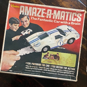 May include: A vintage box for the Amaze-A-Matics game, featuring a white race car with blue stripes and the text "The Fantastic Car with a Brain". The box shows two children playing with the game and includes the text "You Program the Car - You Decide the Action".