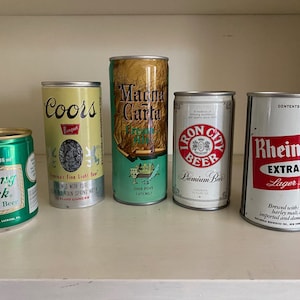 Puede incluir: Seis latas de cerveza de aluminio diferentes, incluyendo Rolling Rock, Coors Banquet, Iron City, Rheingold Extra Dry, y dos más. Las latas están dispuestas en una fila en un estante blanco.