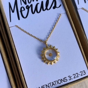 May include: Gold necklace with a clear sun pendant containing a small gold seed. The pendant has a starburst design. The necklace is in a box with the words "New Mercies" and "LAMENTATIONS 3:22-23" printed on it.