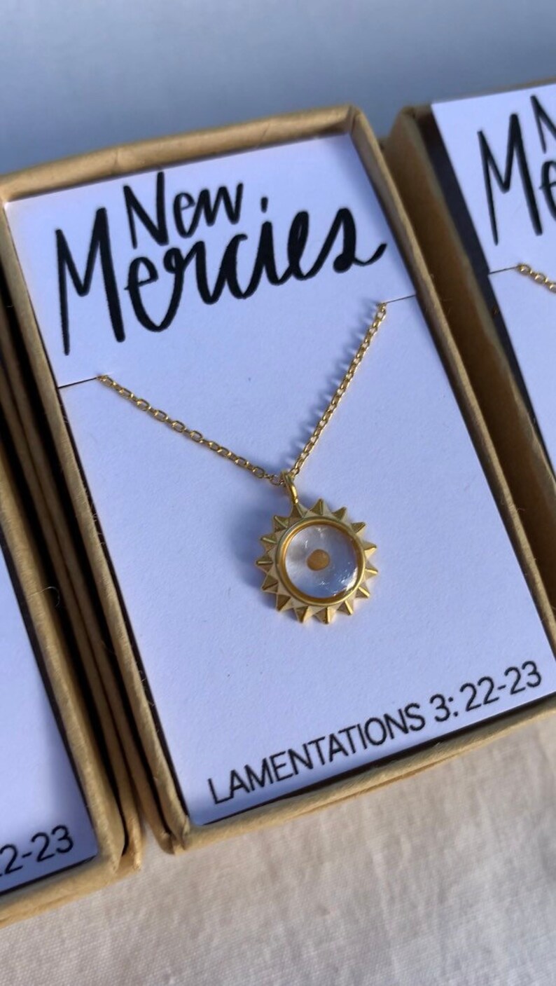May include: Gold necklace with a clear sun pendant containing a small gold seed. The pendant has a starburst design. The necklace is in a box with the words "New Mercies" and "LAMENTATIONS 3:22-23" printed on it.