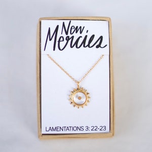May include: A gold necklace with a small sun pendant containing a tiny seed. The necklace is in a box with the words "New Mercies" and "Lamentations 3:22-23" printed on it.