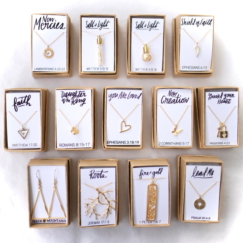 May include: A collection of small, brown boxes with gold necklaces and earrings inside. Each box has a different inspirational quote and scripture reference. The quotes include "New Mercies", "Salt's Light", "Shield of Faith", "Faith", "Daughter of the King", "You Are Loved", "New Creation", "Guard Your Heart", "Roots", "Fire & Gold", and "Lead Me".