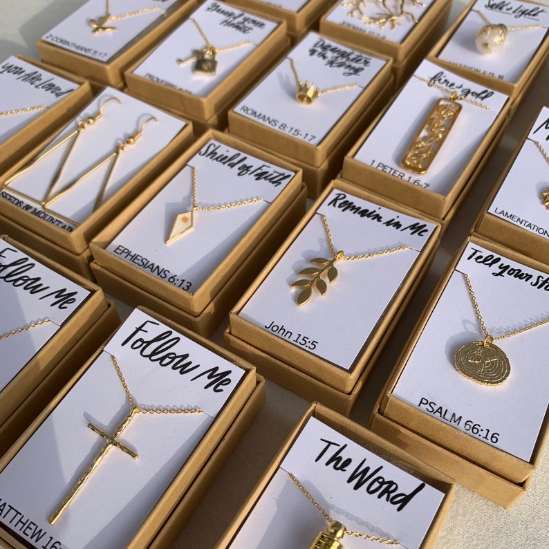 May include: A collection of small, gold-toned necklaces and earrings in individual boxes. Each box has a white background with a different Bible verse and a small, gold-toned pendant. The pendants include a cross, a leaf, a shield, a crown, and a small, round design.