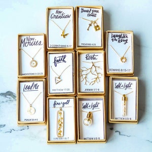 May include: A collection of 12 gold necklaces in small boxes with inspirational quotes and bible verses. Each necklace features a different pendant, including a butterfly, a key and lock, a sun, a triangle, a tree branch, a circle, a compass, a light bulb, and a small vial filled with gold flakes.