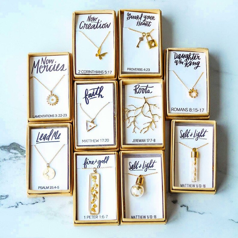 May include: A collection of 12 gold necklaces in small boxes with inspirational quotes and bible verses. Each necklace features a different pendant, including a butterfly, a key and lock, a sun, a triangle, a tree branch, a circle, a compass, a light bulb, and a small vial filled with gold flakes.