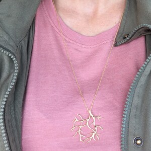 May include: A gold necklace with a delicate tree branch pendant. The necklace is worn by a person wearing a pink t-shirt and a green jacket.