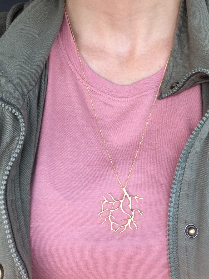 May include: A gold necklace with a delicate tree branch pendant. The necklace is worn by a person wearing a pink t-shirt and a green jacket.