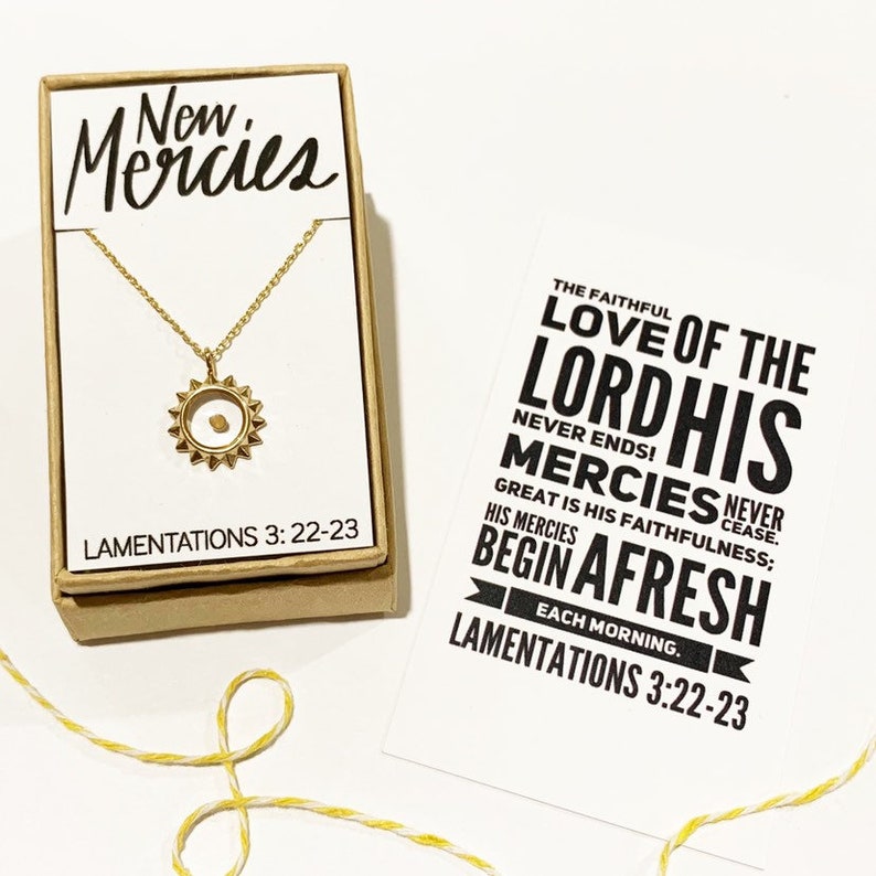 May include: A gold necklace with a small sun pendant containing a tiny seed. The necklace comes in a brown box with the words "New Mercies" and the scripture "Lamentations 3:22-23". A separate card features the scripture "The faithful love of the Lord His mercies never cease. Great is His faithfulness; His mercies begin afresh each morning. Lamentations 3:22-23".
